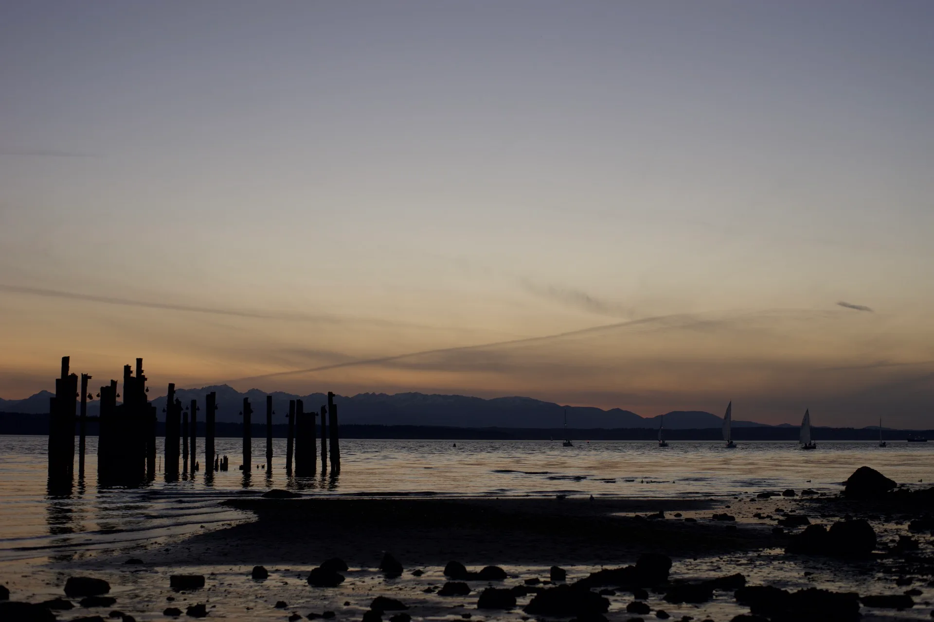 Sunset silhouette of old pier pilings in Puget Sound, sailboats and Olympic Mountains in the distance, low tide exposing rocky shore