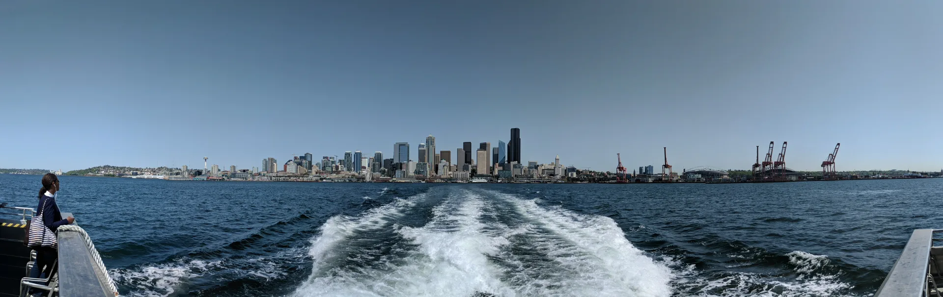 Panorama from a ferry looking back at the Seattle skyline, wake churning behind the boat, Space Needle on the left, port cranes on the right