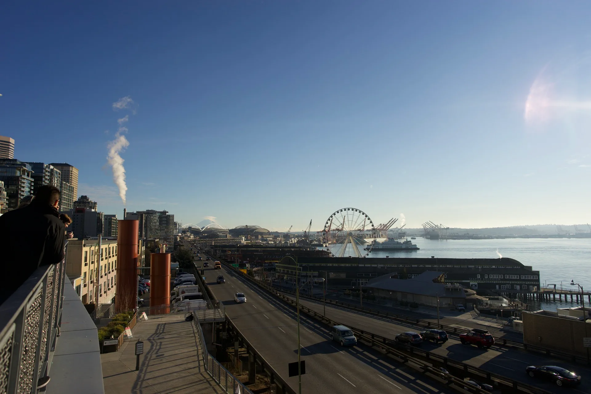 Wide view from elevated position showing the Alaskan Way Viaduct, Seattle Great Wheel, steam rising from a building, someone photographing at the edge of the frame