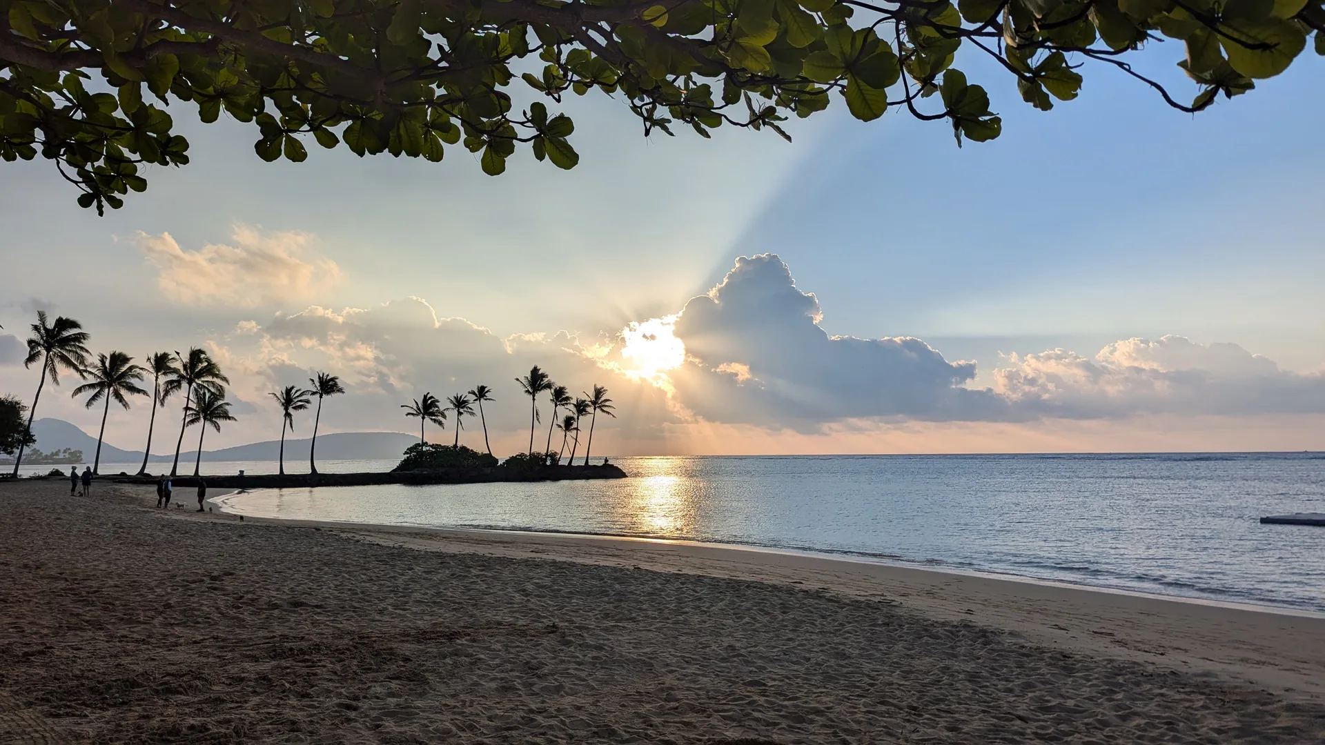 Sunset over a calm lagoon, crepuscular rays bursting from behind clouds, palm trees silhouetted on a small peninsula, framed by overhanging leaves