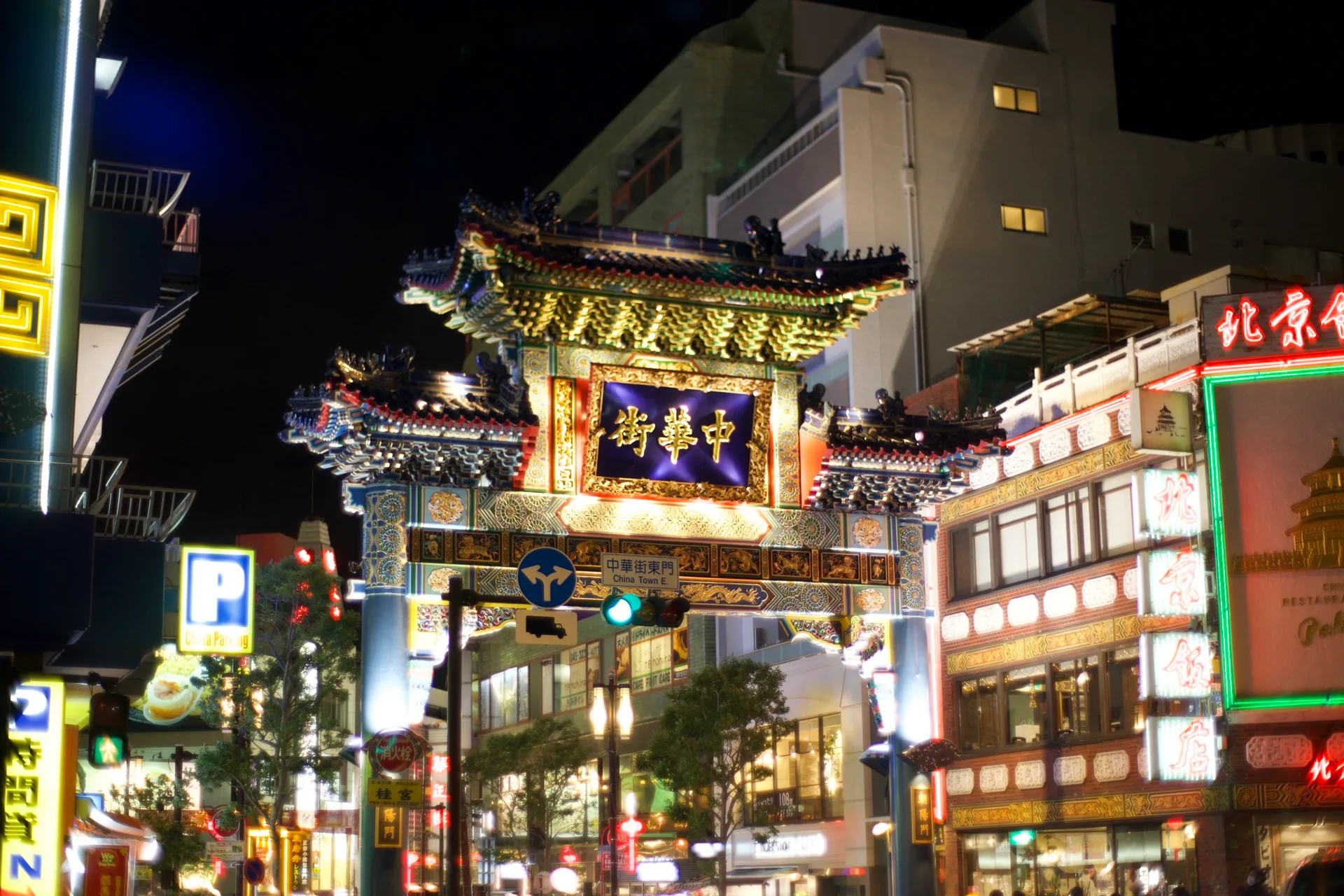 Yokohama Chinatown gate lit up at night, ornate golden Chinese arch with neon restaurant signs glowing around it