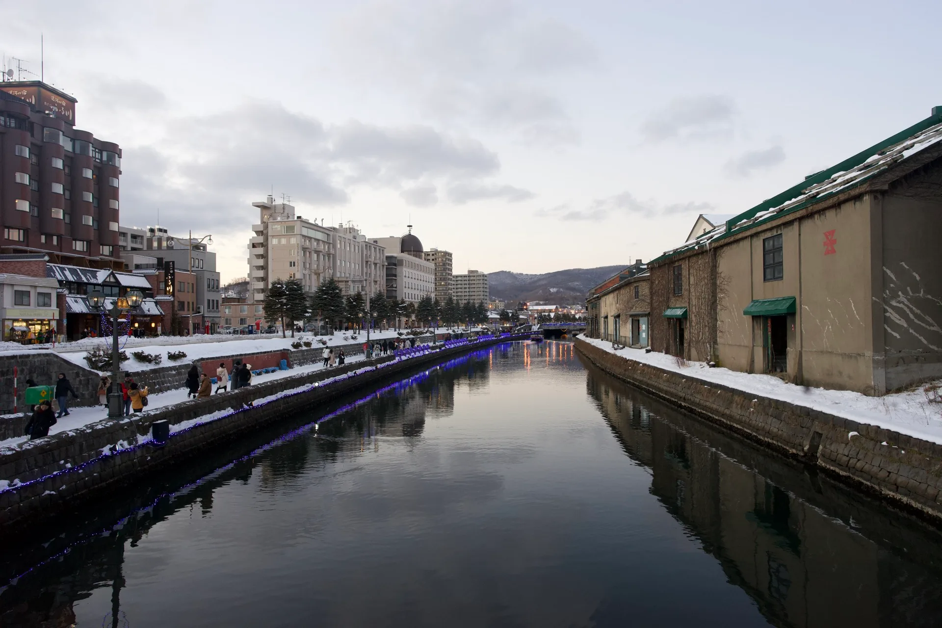 Otaru Canal in winter, snow on the walkways, blue LED lights reflected in perfectly still water, old stone warehouses lining the right bank