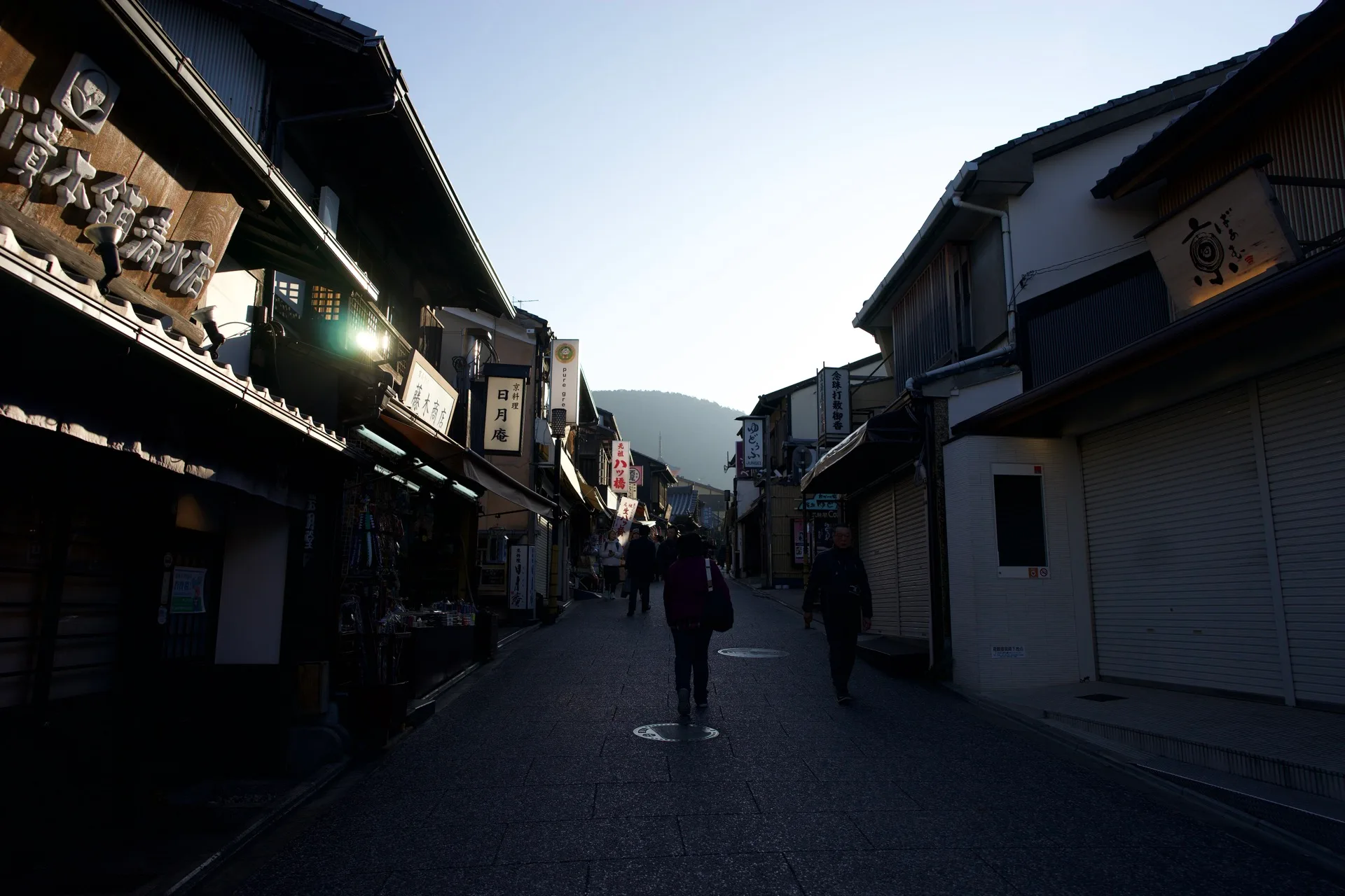 Traditional Japanese shopping street at dusk, low wooden buildings with cloth banners and kanji signs, a lone figure walking ahead into soft backlight