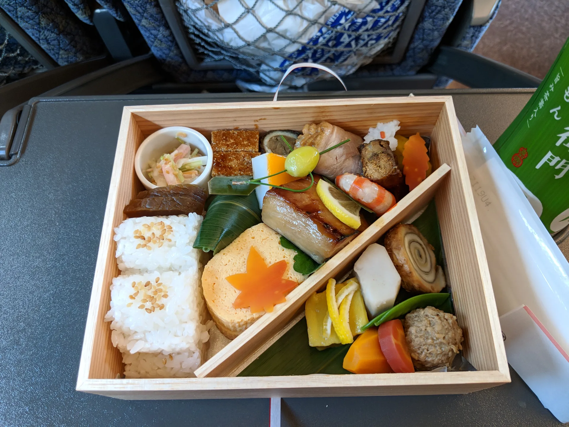 Autumn ekiben in a wooden box on a Shinkansen tray table, compartments of rice, grilled fish, seasonal vegetables with a maple leaf carrot cutout, green tea beside it