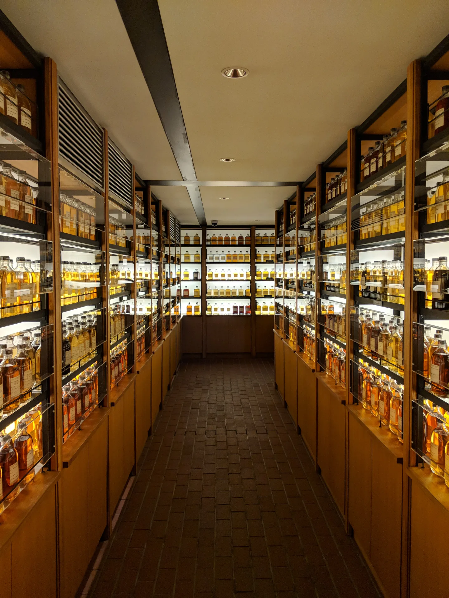 Long corridor of backlit whisky bottles in glass display cases, amber tones glowing warmly, brick floor leading to the vanishing point