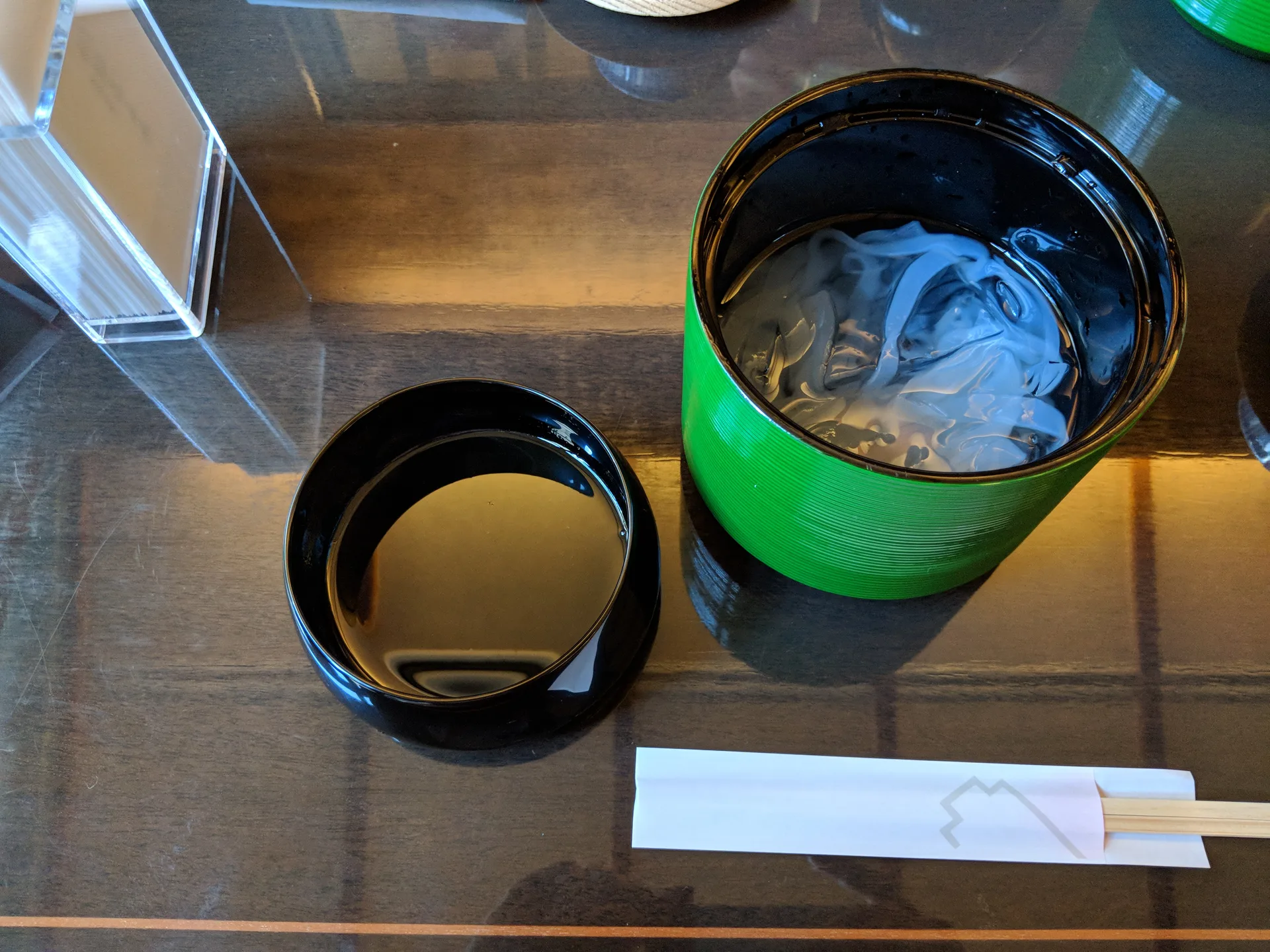 Traditional Japanese sweet in a green lacquerware container with ice, black lacquer cup alongside, wrapped chopsticks on a wooden table